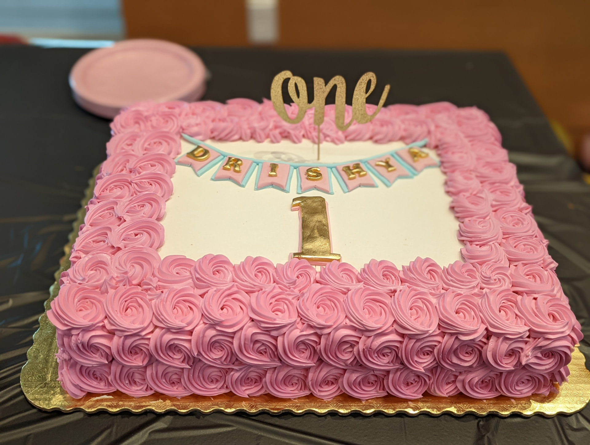 A pink rosette first birthday cake with a gold ONE topper and a name banner